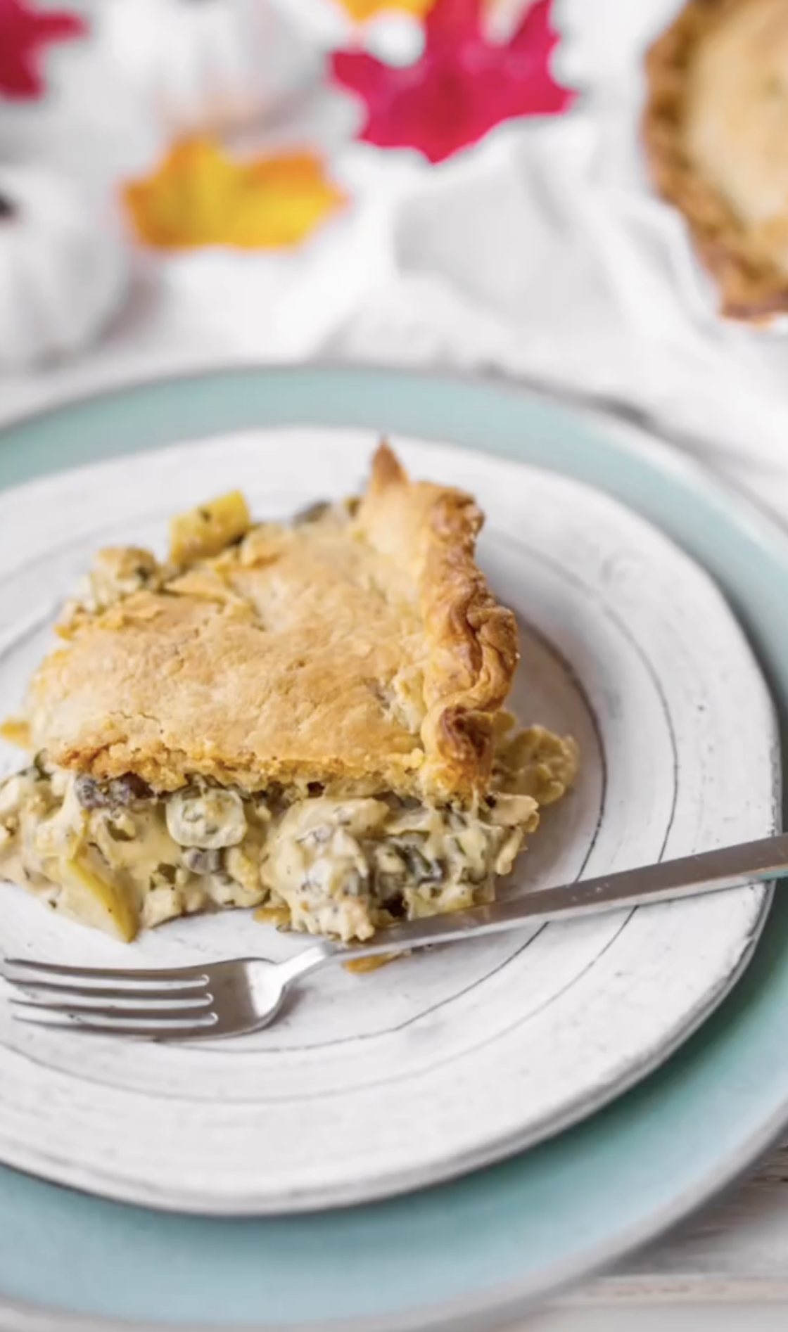 A slice of golden-crusted vegan pie with chunky, creamy filling sits on a white plate with a fork, atop a turquoise-rimmed plate; red and yellow leaf decorations blur in the background.