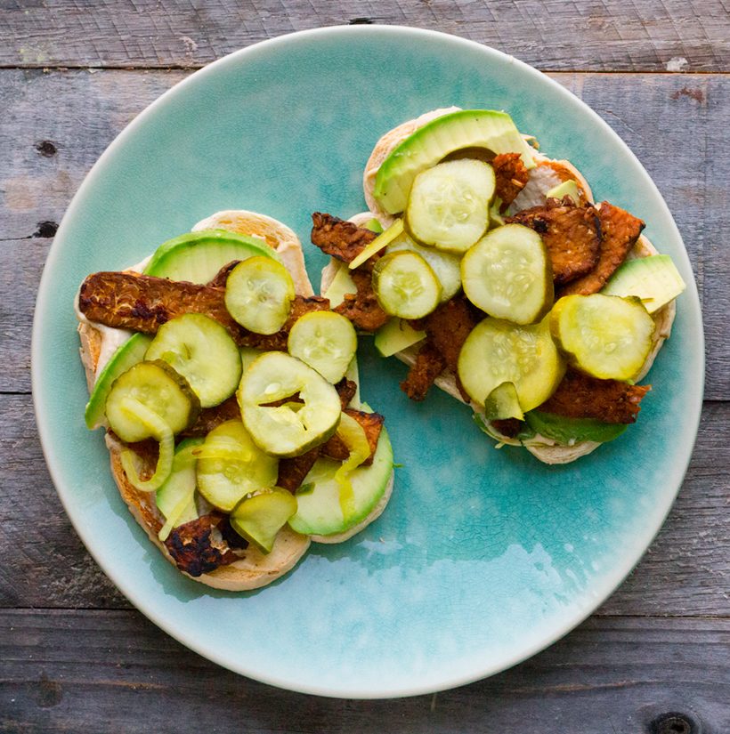 Sourdough toast topped with hummus, avocado, tempeh bacon, and sweet bread and butter pickles.
