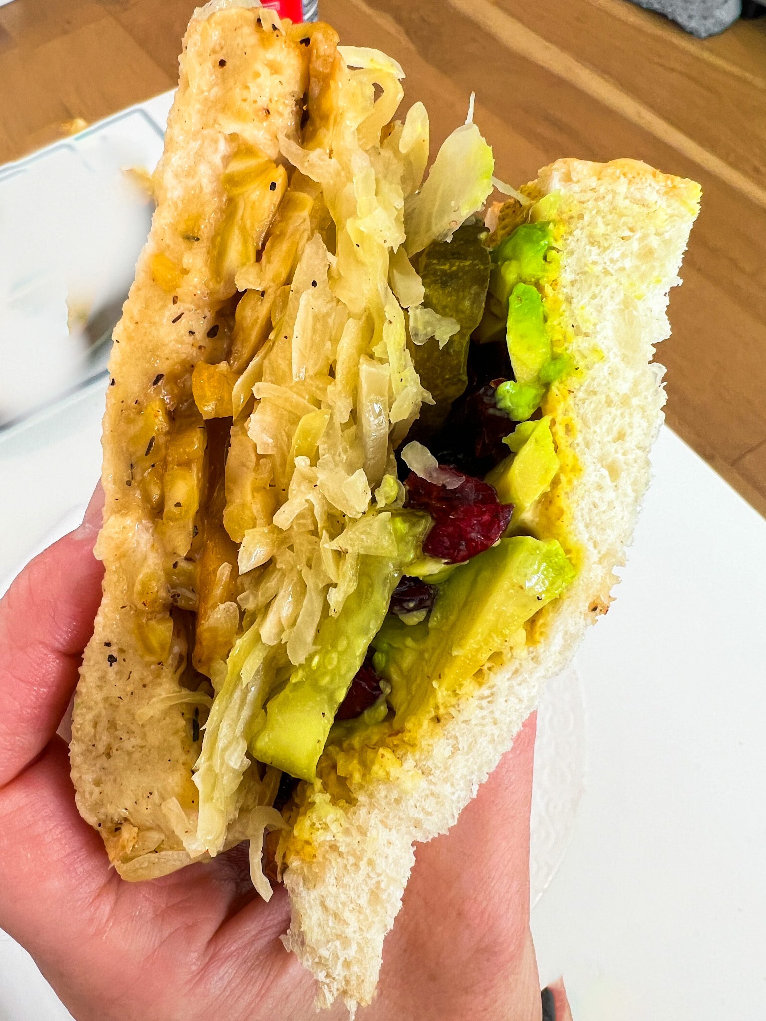 Vegan tempeh Reuben with avocado, sauerkraut, pickles, dried cranberries, mustard, and creamy sauce on toasted sourdough.