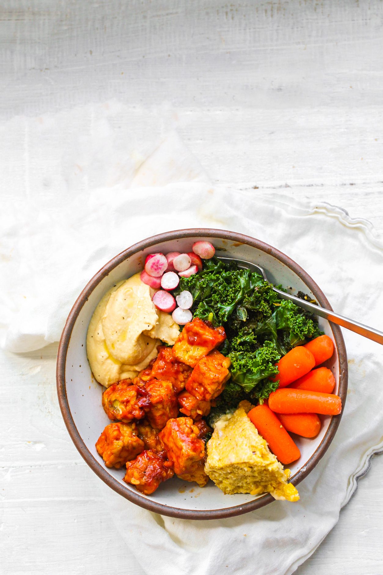 BBQ tempeh cubes over wilted kale with hummus, crunchy carrots and a slice of vegan cornbread.