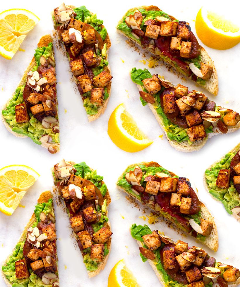 Avocado toast topped with smoky-sweet tofu cubes, heirloom tomatoes, candied almonds, and lemon zest.