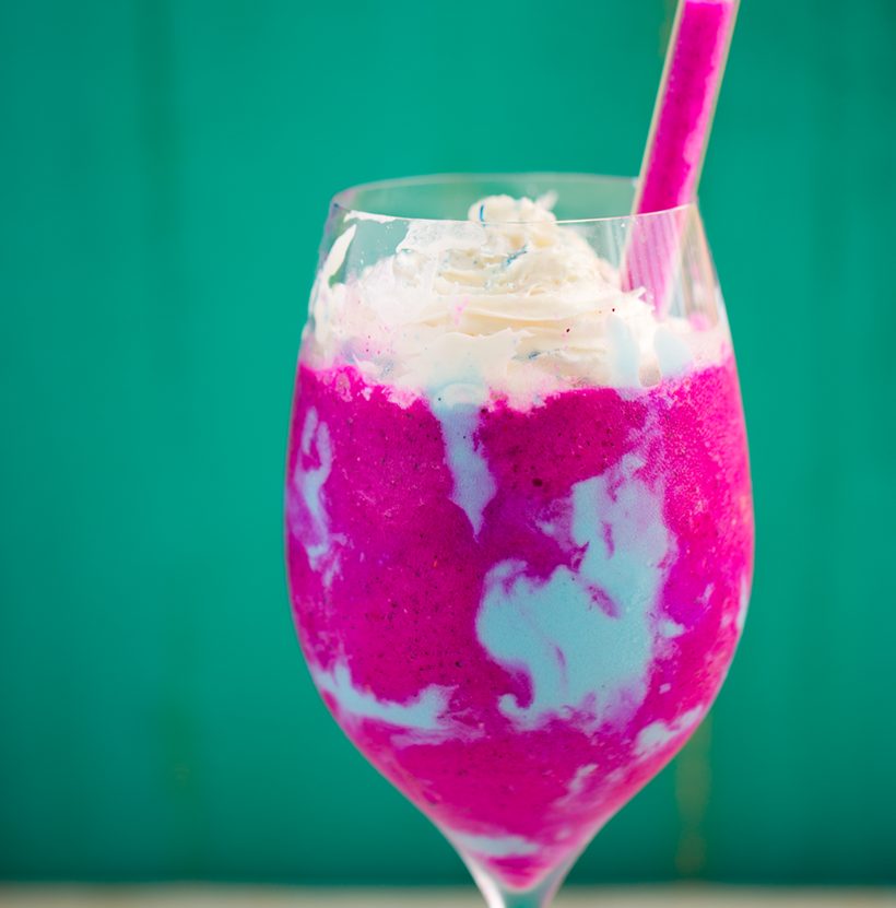 A pink fruit frapp with blue swirls, topped with vegan whipped cream and a dusting of blue powder.