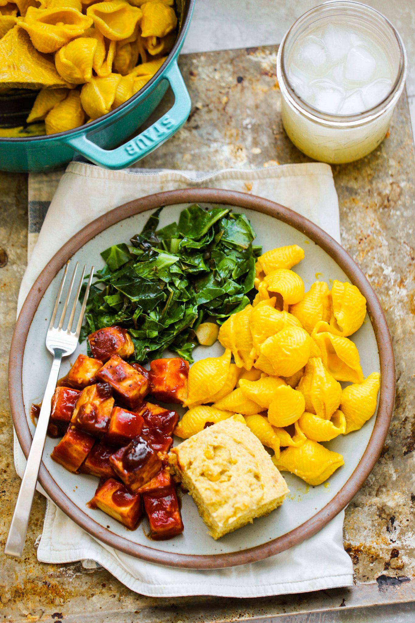 A Southern-style vegan plate with sticky BBQ tofu cubes, cheesy pasta shells, garlicky collard greens and fluffy cornbread.
