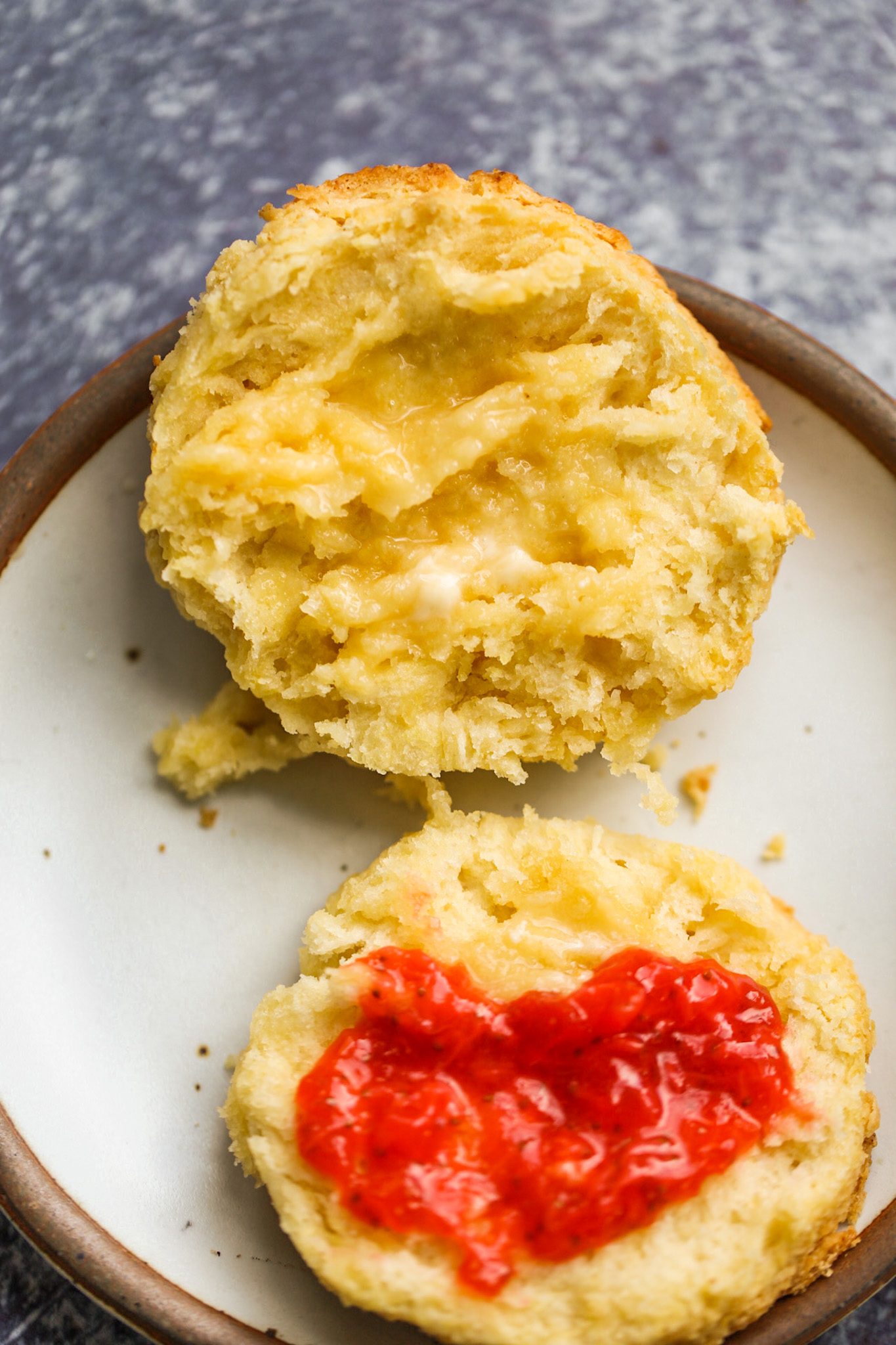 A split vegan buttermilk biscuit with a fluffy, flaky interior, topped with vegan butter and strawberry jam.