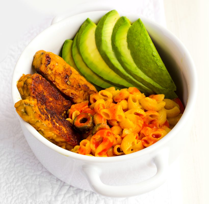 Creamy vegan mac and cheese drizzled with hot sauce, served with avocado and savory-sweet tempeh sticks.